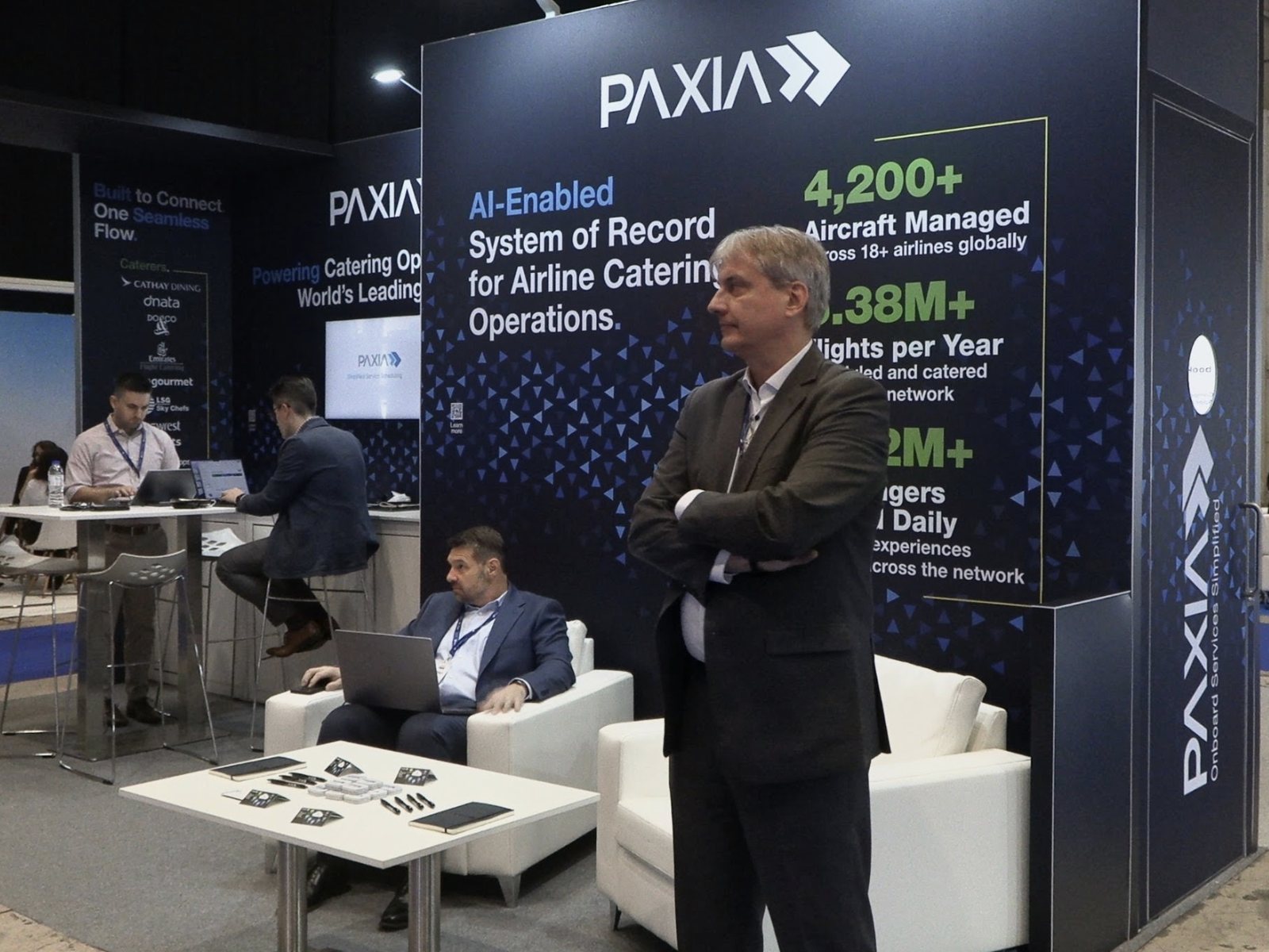 Henning Bruns stands with arms folded at the Paxia booth at World Aviation Festival 2026 while Rodney Duty, Ryan Gaus, and Anthony Harmond work on laptops behind him.